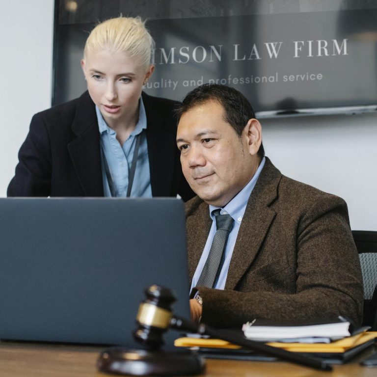 Smart mature ethnic judge in suit working on laptop and discussing details of new case with young female assistant in law firm